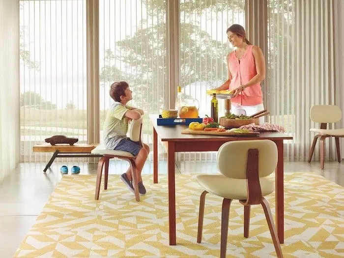 A mother preparing food at a dining table while her young child sits nearby watching in a bright modern room with large windows and vertical blinds.
